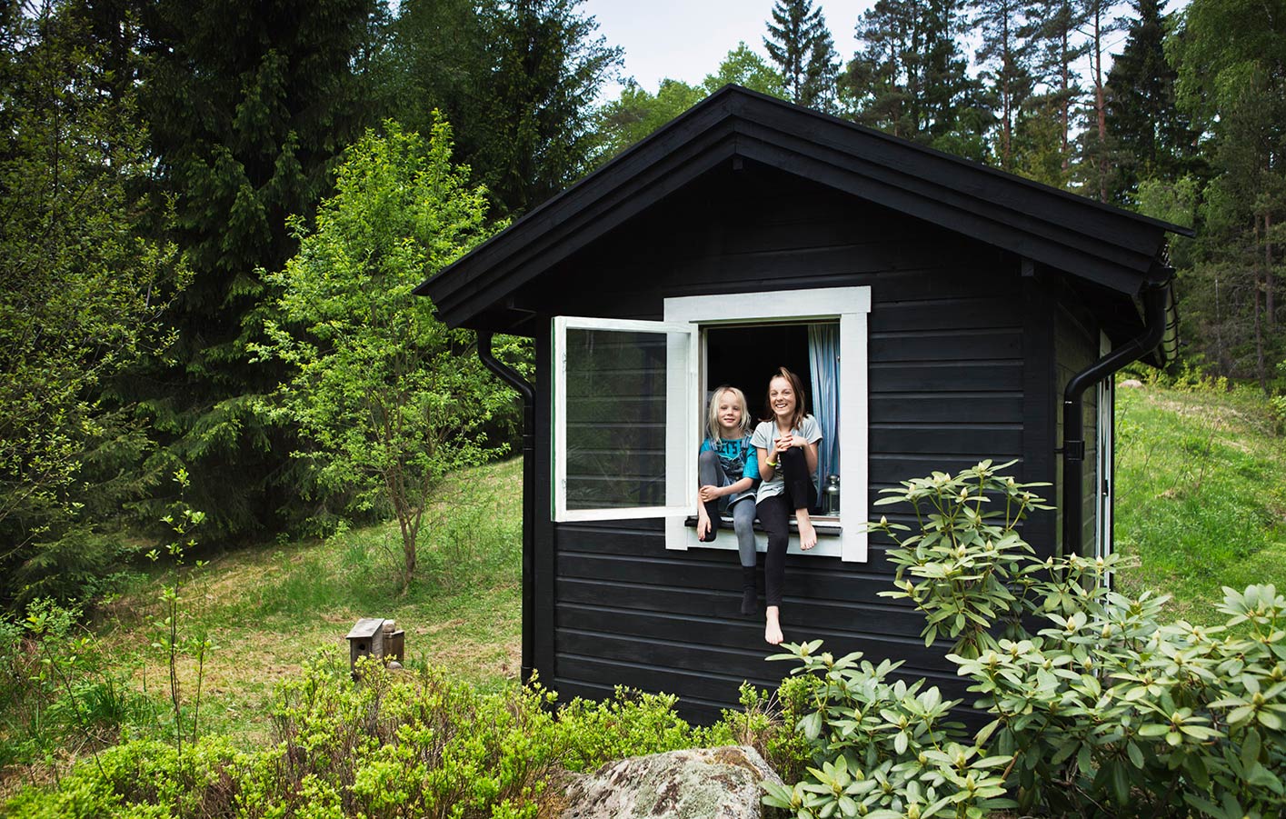 Individuelle Gartenhäuser inkl. Service und Beratung + Aufbau finden Sie bei uns Schwarzes Gartenhaus mitten im Grünen - 2 Mädchnen sitzen auf der Fensterbank